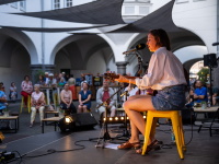 Eine junge Frau spielt Gitarre und singt auf einer Bühne in einem offenen Innenhof, umgeben von aufmerksam lauschendem Publikum.