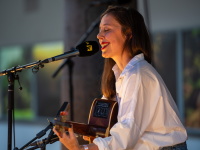 Die Musikerin singt mit geschlossenen Augen ins Mikrofon, begleitet von einer Gitarre, im warmen Bühnenlicht.