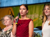 Eine Rednerin in rotem Oberteil spricht auf der Bühne, links und rechts neben ihr stehen zwei weitere Frauen vor einem Banner.