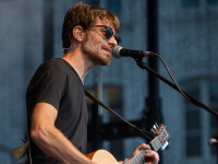 Ein Musiker mit Akustikgitarre singt leidenschaftlich ins Mikrofon. Er trägt Sonnenbrille und schwarze Kleidung.