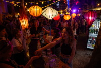 Eine junge Frau reicht einem Gast an der festlich geschmückten Outdoor-Bar ein Getränk. Über ihr leuchten orangefarbene und rote Lampions.