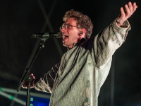 Ein junger Mann mit Brille und Ohrstöpseln singt laut mit weit geöffneten Armen auf der Bühne.