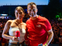 Eine Moderatorin in weißem Top neben deinem Mann im roten T-Shirt, im Hintergund das Publikum