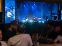 Kamrad steht im Spotlight mit Gitarre vor riesiger Leinwand. Die Bühne ist in tiefblaues Licht getaucht, das Publikum verfolgt gespannt seinen Auftritt.