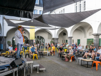 Blick in den Innenhof der Stadtgalerie mit Sonnensegeln über orange und gelben Stühlen. Links moderiert eine Person auf der Bühne. 