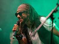 er Sänger lächelt ins Mikrofon auf der Bühne. Er trägt eine Sonnenbrille, Dreadlocks, Stirnband und eine weiße T-Shirt-Weste-Kombination. Die grüne Bühnenbeleuchtung taucht ihn in ein atmosphärisches Licht.
