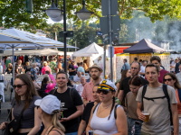 Die Menschen schlendern vorbei an Marktstände, einige mit Getränken in der Hand. Im Hintergrund: weiße Marktzelte und Verkaufsstände. 
