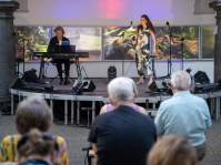 Sängerin in bodenlangem, floralem Kleid singt ins Mikrofon, während ein Pianist im Hintergrund spielt. Das Publikum sitzt nahe an der Bühne in einem Arkadenhof.