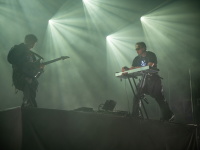 Zwei Musiker von „Two Colors“ in dunkler Kleidung auf erhöhter Bühne, umgeben von intensivem Lichtstrahleneffekt. Links ein Gitarrist, rechts ein Keyboarder mit leuchtendem Ankersymbol auf dem Shirt.