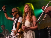 Mitglieder von „Oku & the Reggaerockers“ spielen energiegeladen auf der Bühne. Links der Sänger mit Dreadlocks und erhobenem Arm, rechts eine Saxophonistin in weißem Sommerkleid. Die Bühne ist in grünes Licht getaucht.
