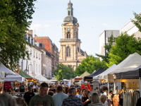 Menschen schlendern durch eine belebte Marktstraße mit Verkaufsständen. Im Hintergrund erhebt sich die Basilika St. Johann, eingerahmt von alten Stadtgebäuden und sommerlichem Grün.
