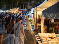 Marktgasse in der Dämmerung mit bunten Ständen und leuchtender Auslage. Familien, Kinder und Paare schlendern durch die beleuchtete Szenerie unter hohen Bäumen.