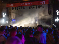 Große Menschenmenge vor der SR1-Bühne bei Nacht. Zwei Musiker auf einem Podest, umgeben von Nebel und eindrucksvollen Lichtstrahlen, mit großflächigen Schwarz-Weiß-Projektionen auf LED-Wänden.