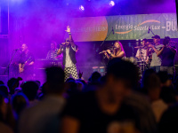 Die Band Oku & the Reggaerockers mit kompletter Besetzung im violetten Bühnenlicht. Im Vordergrund der Sänger mit erhobener Hand, flankiert von Gitarrist, Keyboarder, Flötistin, Trompeter und Drummer.