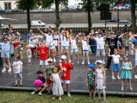 ine große Gruppe von Kindern und Jugendlichen tanzt gemeinsam auf einer Bühne unter freiem Himmel, während zahlreiche Zuschauer am Bühnenrand mitfiebern und applaudieren.