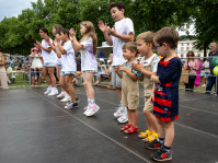 Kinder verschiedener Altersstufen stehen nebeneinander auf einer Bühne und klatschen rhythmisch bei einer Tanzvorführung. Dahinter verfolgen Eltern und Gäste das Geschehen.