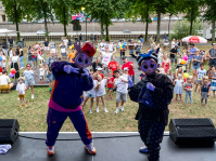 Zwei bunte Maskottchen posieren auf der Bühne vor einer großen Kindergruppe, die fröhlich in die Kamera winkt. Bunte Luftballons und lachende Gesichter prägen das Bild.