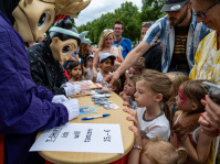 Kinder stehen Schlange und reichen zwei Maskottchen Flyer zum Signieren. Die Stimmung ist gespannt und neugierig – es wird gelacht, beobachtet und gesammelt.