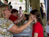 Eine Frau schminkt konzentriert ein Mädchen mit Regenbogenfarben im Gesicht. Im Hintergrund lassen sich weitere Kinder bemalen, während Eltern das Geschehen begleiten.