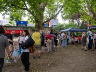amilien, Kinder und Besucher reihen sich unter schattigen Bäumen vor einem Stand auf. 