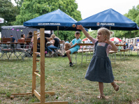 Ein kleines Mädchen mit bemaltem Gesicht wirft lachend einen Ball an einer Schnur auf ein Holzspielgerät. Im Hintergrund sitzen Menschen unter Biergarten-Schirmen und genießen das Fest.