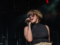 Sängerin singt leidenschaftlich mit Sonnenbrille und schwarzem Top vor dunklem Bühnenhintergrund.
