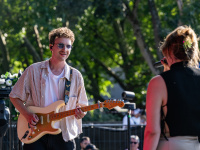 Gitarrist mit Sonnenbrille spielt lächelnd E-Gitarre auf einer Bühne, im Vordergrund steht eine Sängerin.