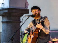 Mann mit Bart und schwarzer Schirmmütze singt auf einer Bühne und spielt Gitarre, im Vordergrund eine steinerne Säule.