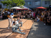 Vier Tänzer performen auf einem Platz vor Zuschauern, einer steht gestikulierend, drei sitzen.