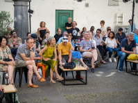 Publikum sitzt auf einem Platz an Tischen und verfolgt aufmerksam ein Konzert im Freien.