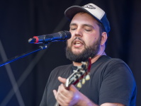 Musiker mit schwarzem Shirt und Kappe singt in ein Mikrofon und spielt dabei eine rote E-Gitarre.