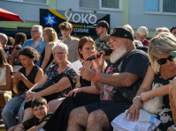 Mann mit Mikrofon und schwarzer Cap liest von einem Zettel ab, umgeben von sitzendem Publikum.