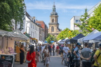 Eine Marktstraße mit vielen Ständen, Besucherinnen und Besuchern führt auf eine Kirche im Hintergrund zu.