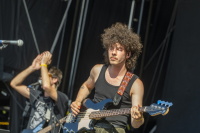 Bassist mit wilder Lockenmähne spielt versunken E-Bass, Bandkollege im Hintergrund unscharf.