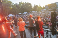 Bläsergruppe spielt vor riesiger Zuschauermenge auf Open-Air-Bühne bei Sonnenuntergang.