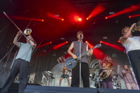 Sänger mit E-Gitarre und Bläsersektion rocken Bühne unter dramatischem rotem Licht.