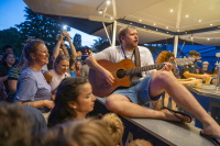 Sänger mit Gitarre performt sitzend auf Bartresen mitten unter lachenden Gästen.