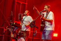 Zwei Musiker mit Gitarre und Mikrofon performen ausgelassen im roten Bühnenlicht.