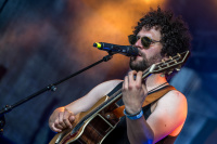 Lockenköpfiger Musiker mit Sonnenbrille singt am Mikrofon und spielt Akustikgitarre, warmes Bühnenlicht im Hintergrund.