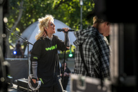 Blonde Sängerin mit Tamburin singt leidenschaftlich ins Mikrofon, im Hintergrund ein Gitarrist im Karohemd, Bäume.