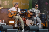 Zwei Musiker sitzen mit Akustikgitarren auf Barhockern, spielen vor Schlagzeug auf Bühne.